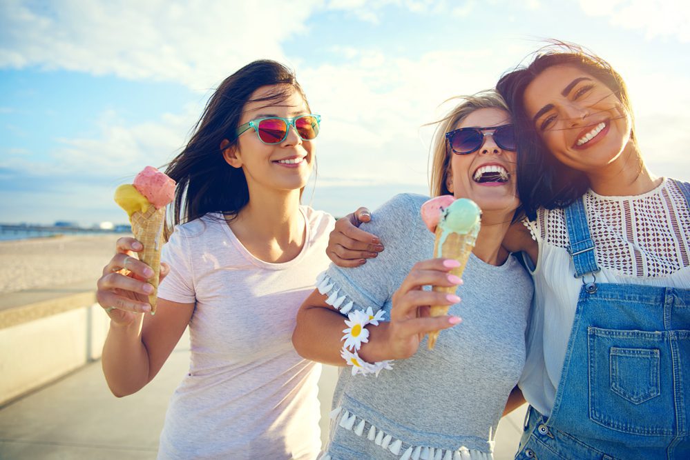 Three young women smile and laugh as they walk near a beach holding ice cream cones