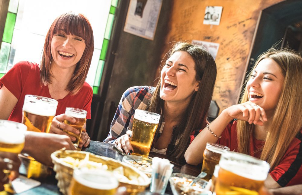 Three women laugh at a restaurant table with glasses of beer