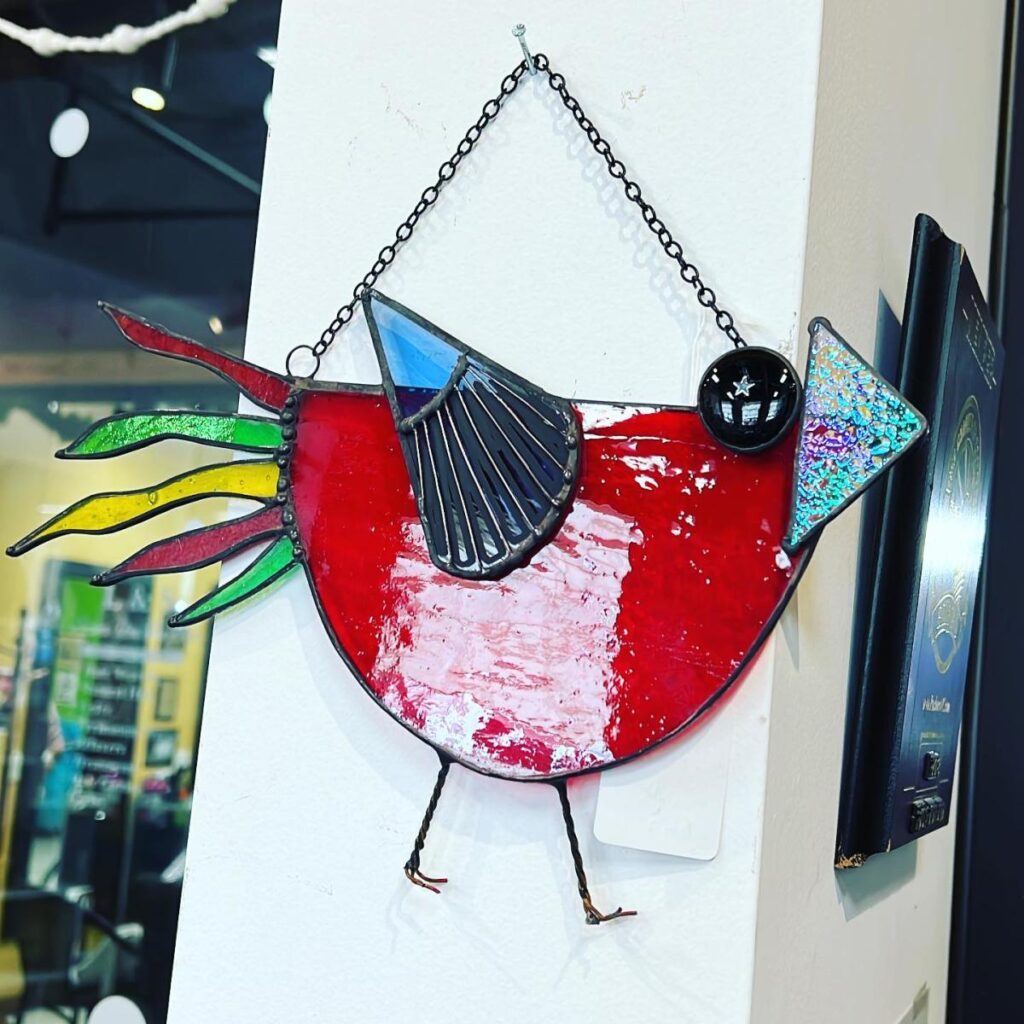 A stained glass bird designed in a modern style hangs from a nail on a white pillar