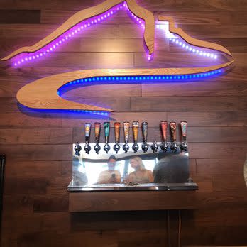 A wooden table with beer taps under neon wall art depicting a mountain