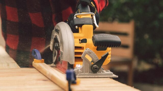 A person wearing a black and red checked shirt uses a power saw on a piece of lumber