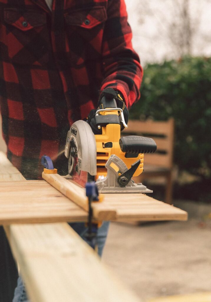 A person wearing a black and red checked shirt uses a power saw on a piece of lumber