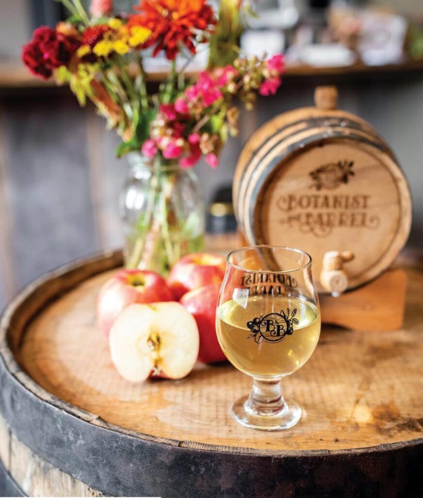A glass of cider presented on top of a barrel with apples and colorful flowers