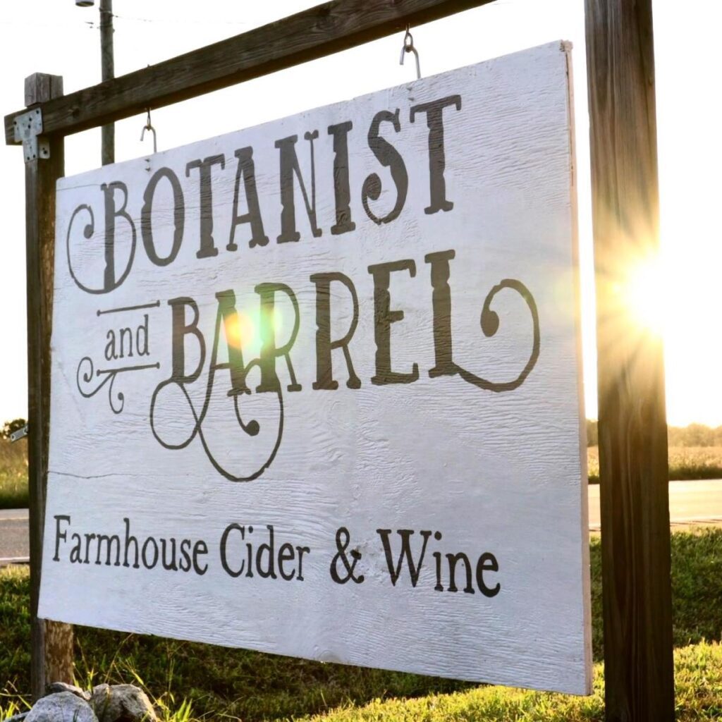 A white painted wooden sign reading Botanist and Barrel, Farmhouse Cider & Wine