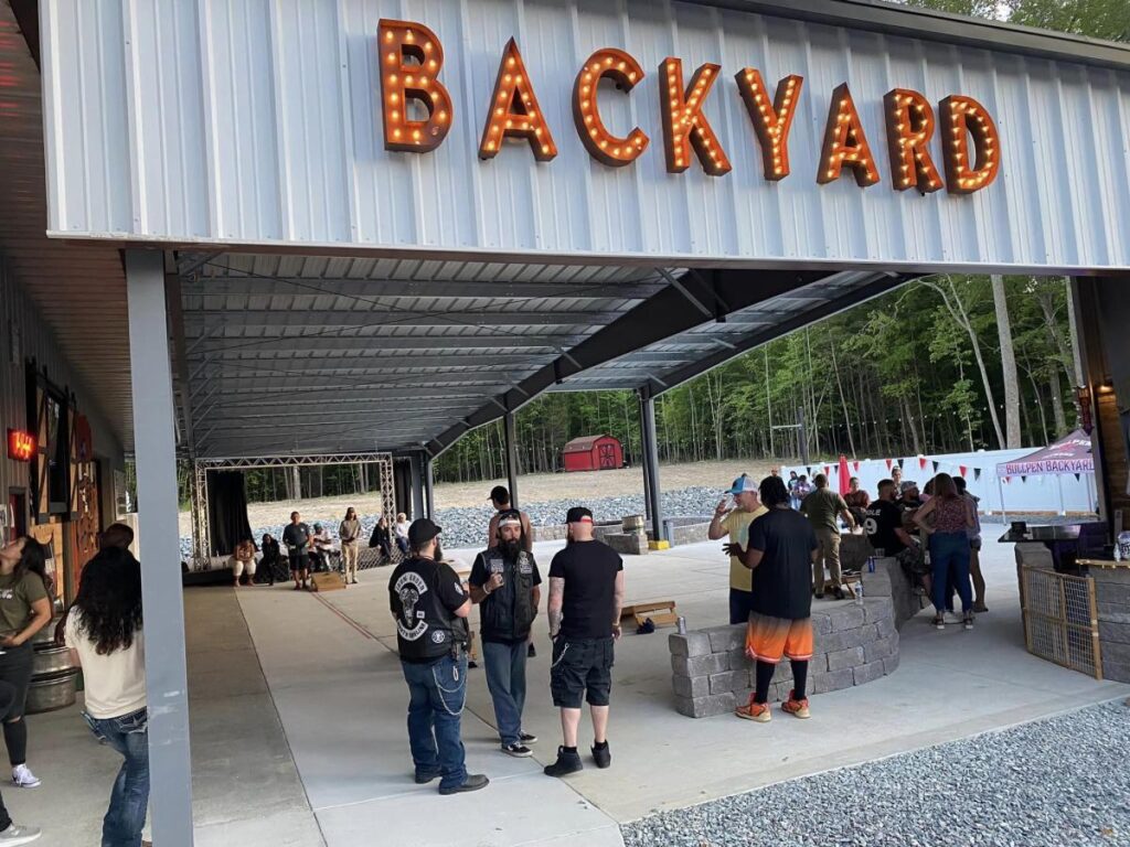 Clusters of people socialize under a metal ramada roof that has a red sign reading Backyard