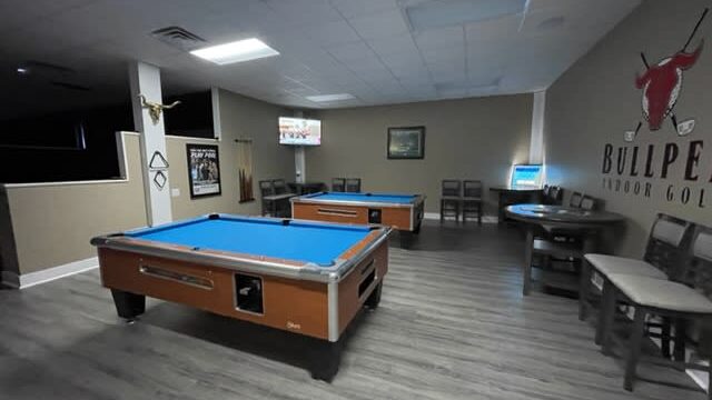 Two blue-topped pool tables in a large room, with tables and chairs along the wall