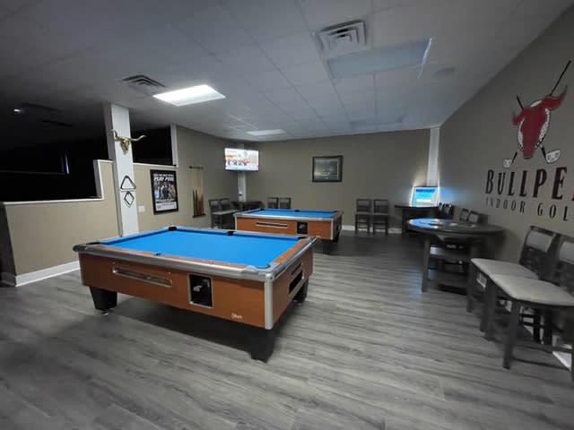 Two blue-topped pool tables in a large room, with tables and chairs along the wall