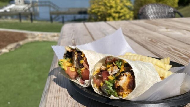 Two halves of a burrito served in a black plastic basket sitting on a wooden table in front of a lake