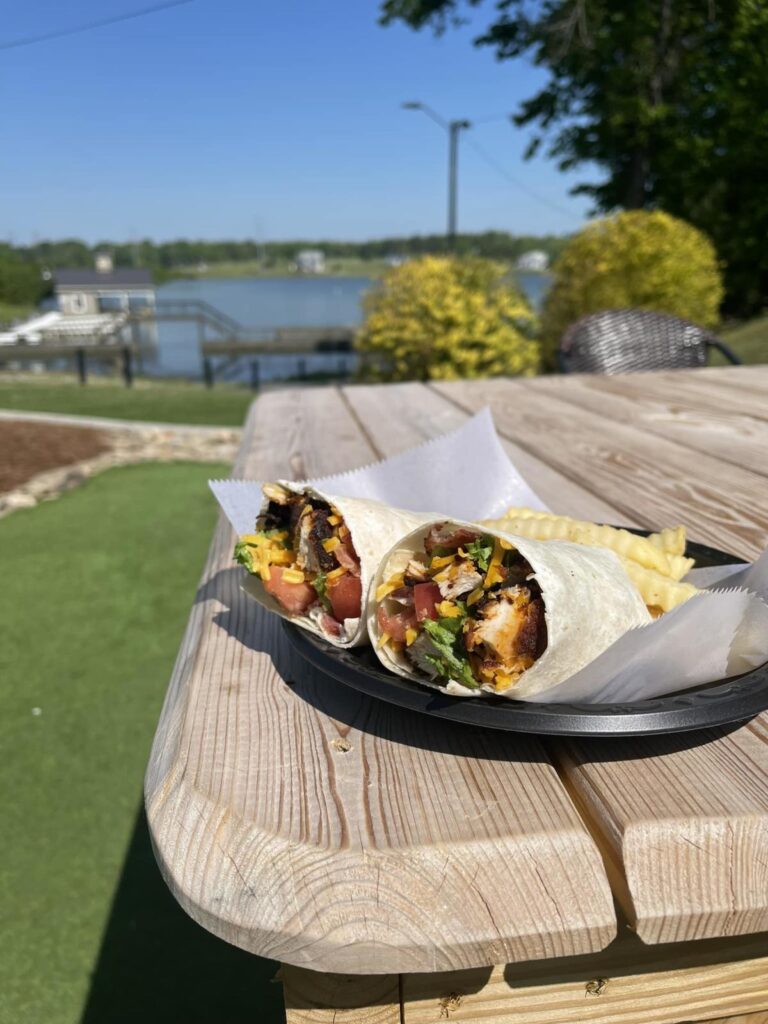 Two halves of a burrito served in a black plastic basket sitting on a wooden table in front of a lake