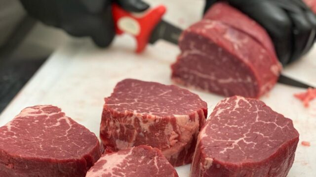 A butcher wearing black plastic gloves slices raw meat on a counter