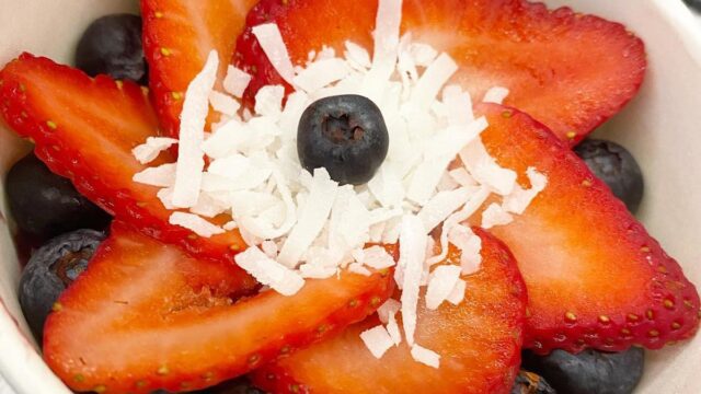 Sliced strawberries fanned out in a flower shape on top of blueberries with coconut flakes and a blueberry on top