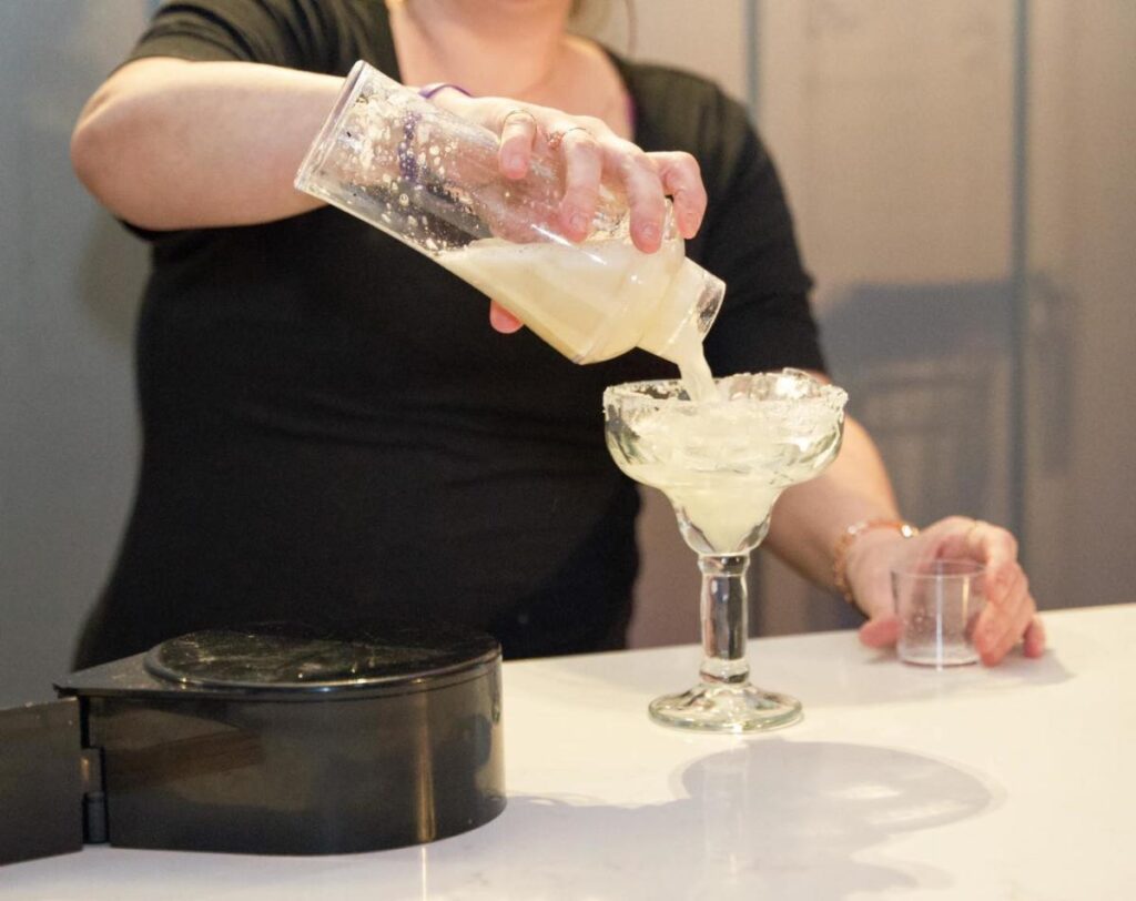 A bartender wearing black pours a margarita into a margarita glass rimmed with salt