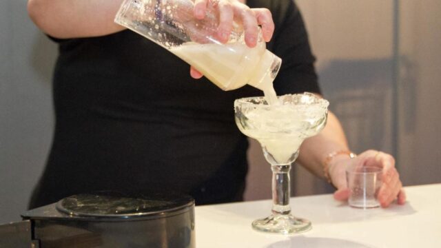 A bartender wearing black pours a margarita into a margarita glass rimmed with salt