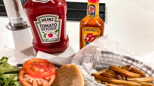 Two black plastic baskets with a cheeseburger and French fries sit on a counter with bottles of ketchup behind