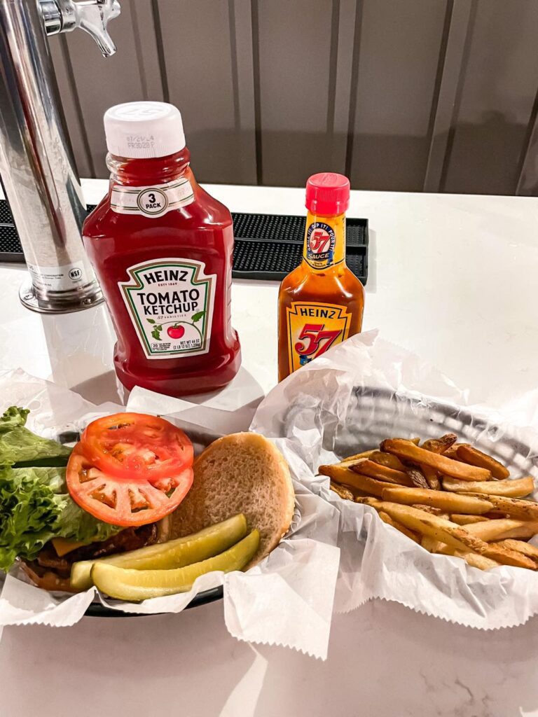 Two black plastic baskets with a cheeseburger and French fries sit on a counter with bottles of ketchup behind