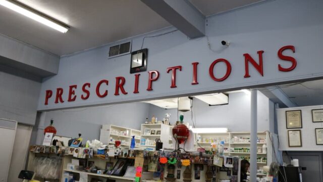 A pharmacy counter inside a store with a large red sign above the counter reading Prescriptions