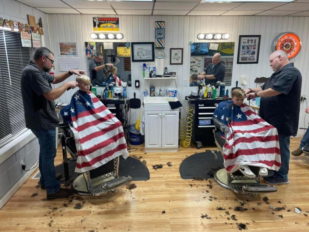 Two young boys getting haircuts side by side at a barbershop, draped in American flag smocks