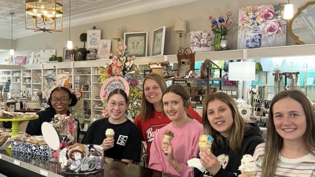 Five young women sit at a counter holding ice cream cones and smiling with an adult woman smiling and standing behind them