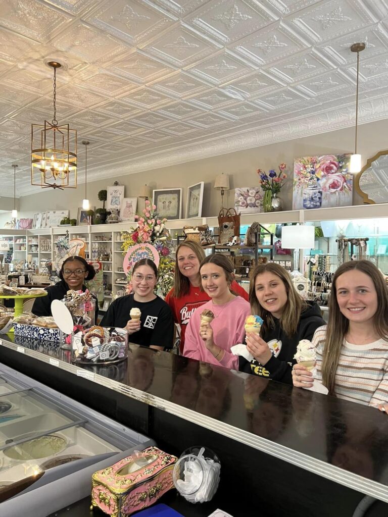 Five young women sit at a counter holding ice cream cones and smiling with an adult woman smiling and standing behind them