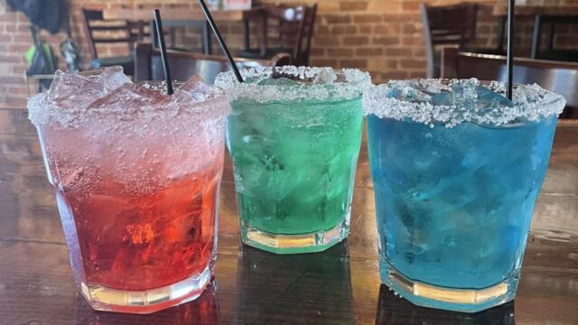 Red, green, and blue drinks with ice cubes and rimmed with salt displayed on top of a bar