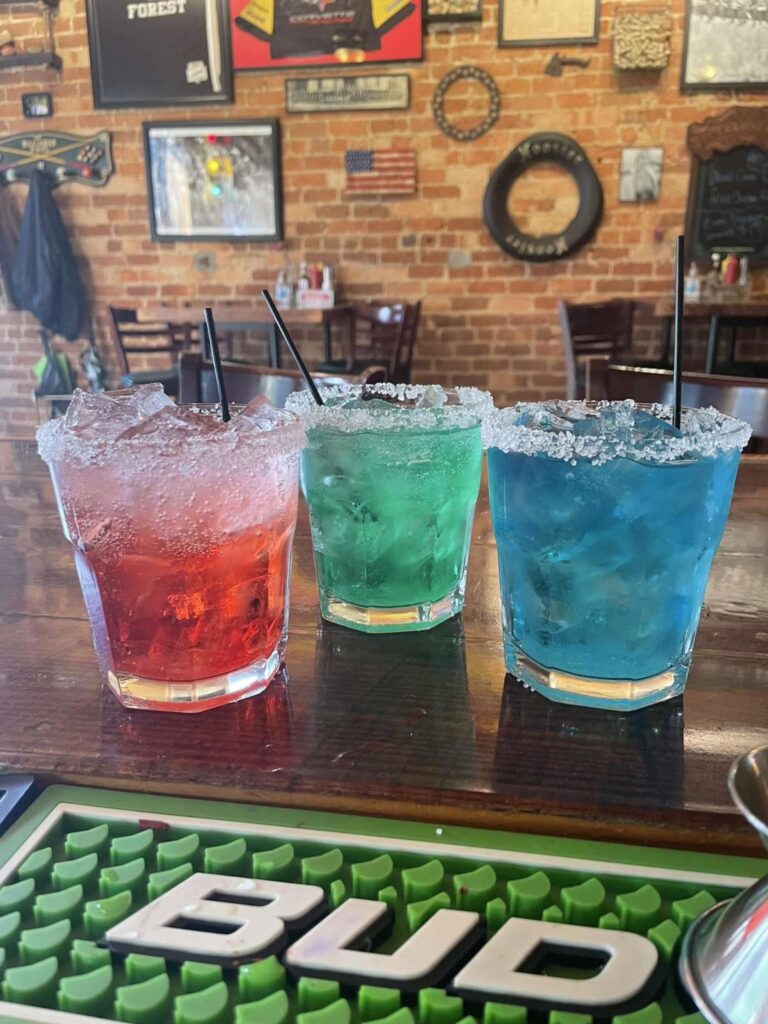 Red, green, and blue drinks with ice cubes and rimmed with salt displayed on top of a bar