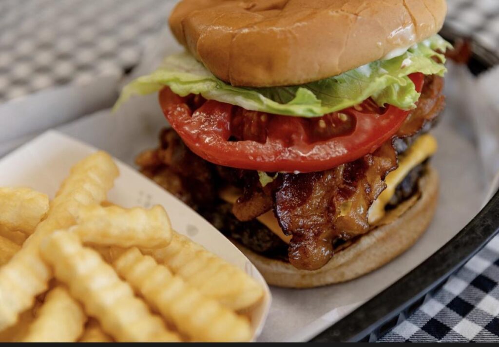 A tall bacon cheeseburger and French fries on a white plate on top of a black and white checked tablecloth
