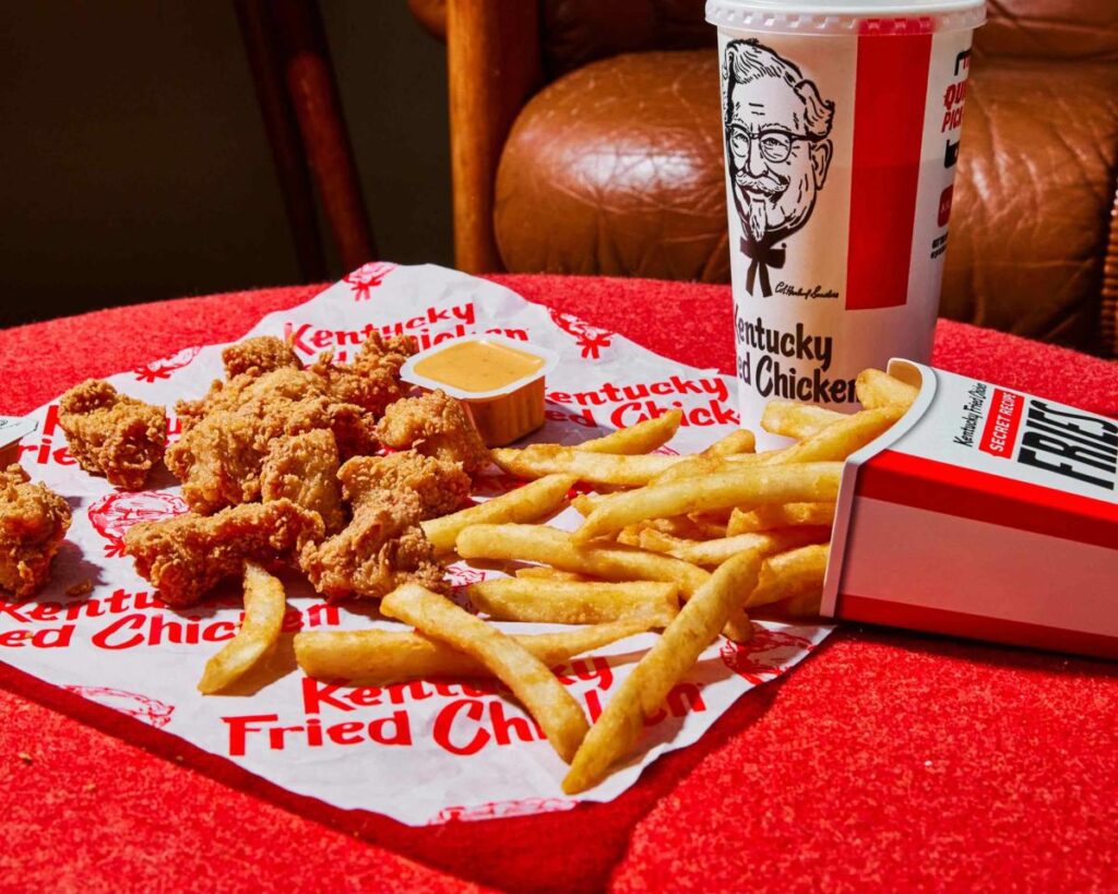 Chicken fingers and French fries spilling out of the fry container onto a white Kentucky Fried Chicken wrapper on a red table