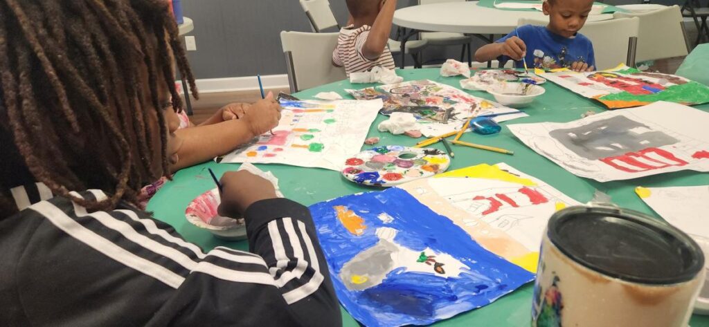 Children sitting at a table painting with paintbrushes and palettes of paint