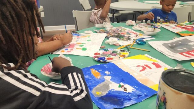 Children sitting at a table painting with paintbrushes and palettes of paint