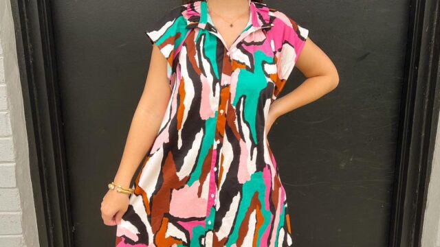 A smiling woman wears a colorful abstract print dress with pink, green, white, and brown