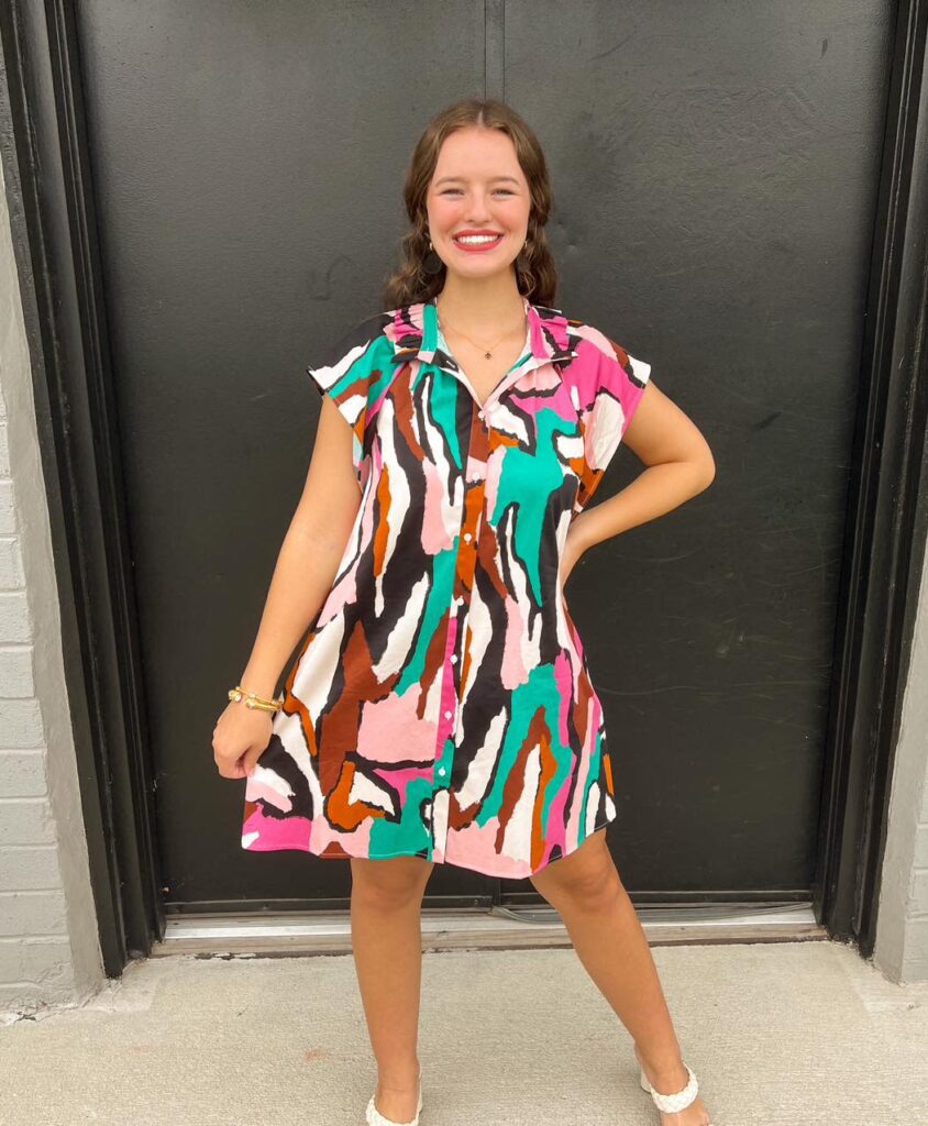 A smiling woman wears a colorful abstract print dress with pink, green, white, and brown