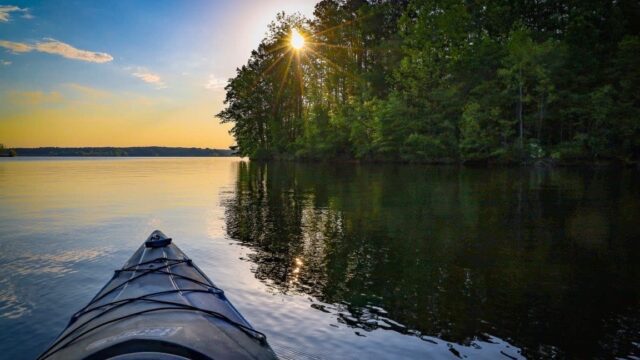 The tip of a kayak on a lake surrounded by green trees