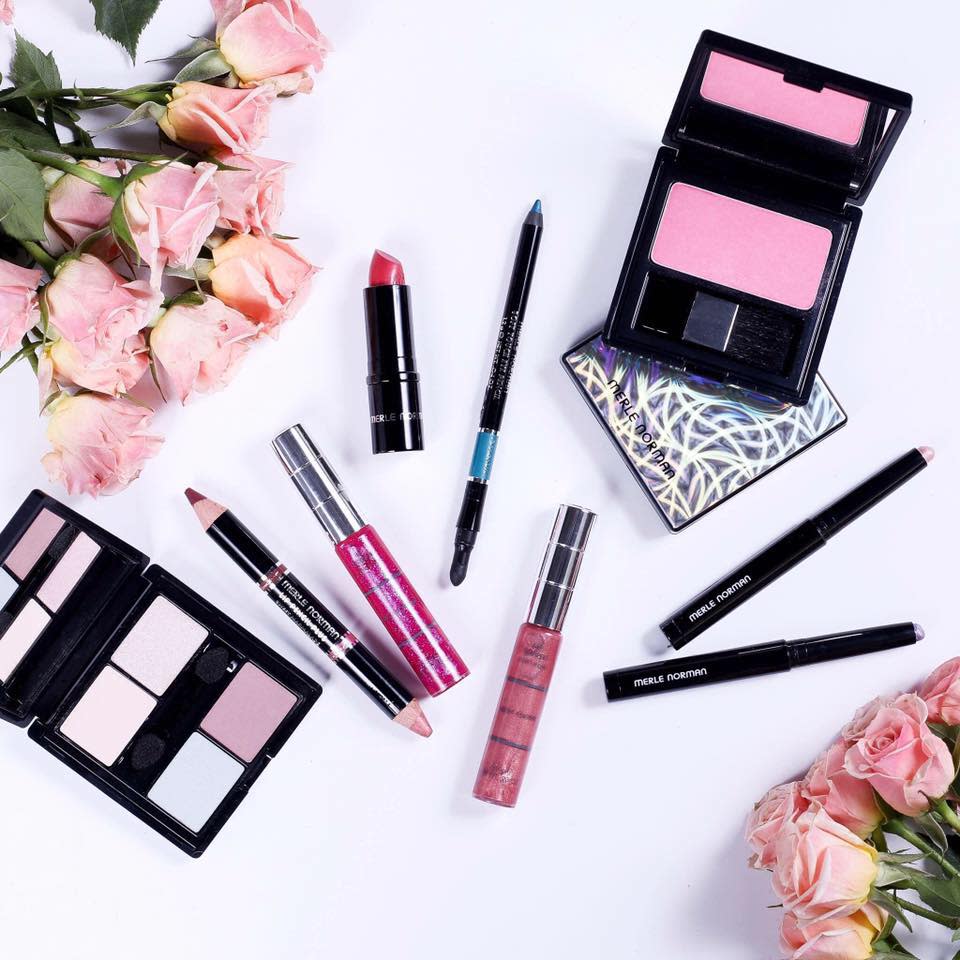 A selection of eye shadow palettes, lipsticks, and eyeliner pencils displayed on a white background with two bouquets of pink roses.