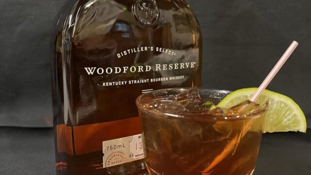 A full bottle of Woodford Reserve whiskey and a glass of whiskey with a lemon wedge placed in front