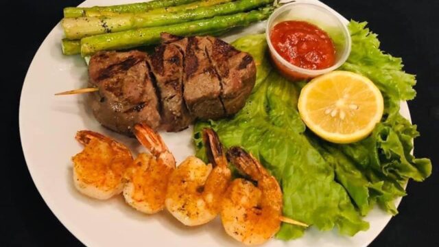 A white plate with grilled meat, four shrimp on a skewer, asparagus, a piece of lettuce, half a lemon, and red sauce in a plastic cup