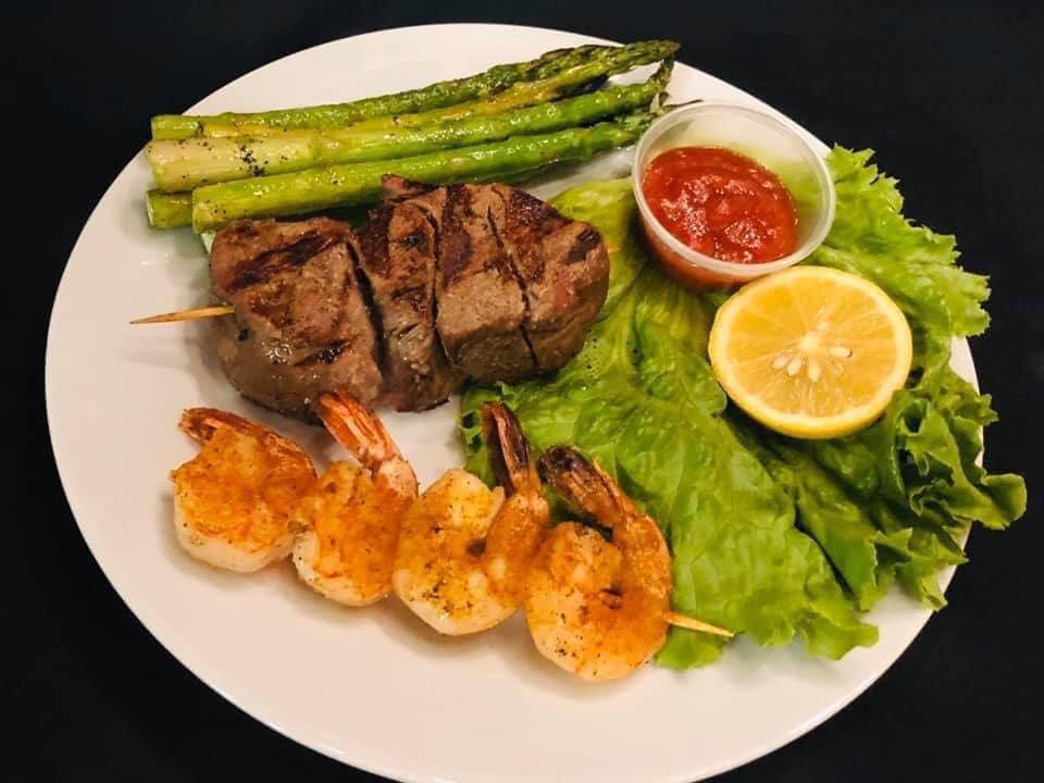 A white plate with grilled meat, four shrimp on a skewer, asparagus, a piece of lettuce, half a lemon, and red sauce in a plastic cup