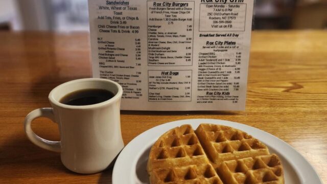 A white mug of coffee and a waffle on a white plate sit on a table in front of a diner menu