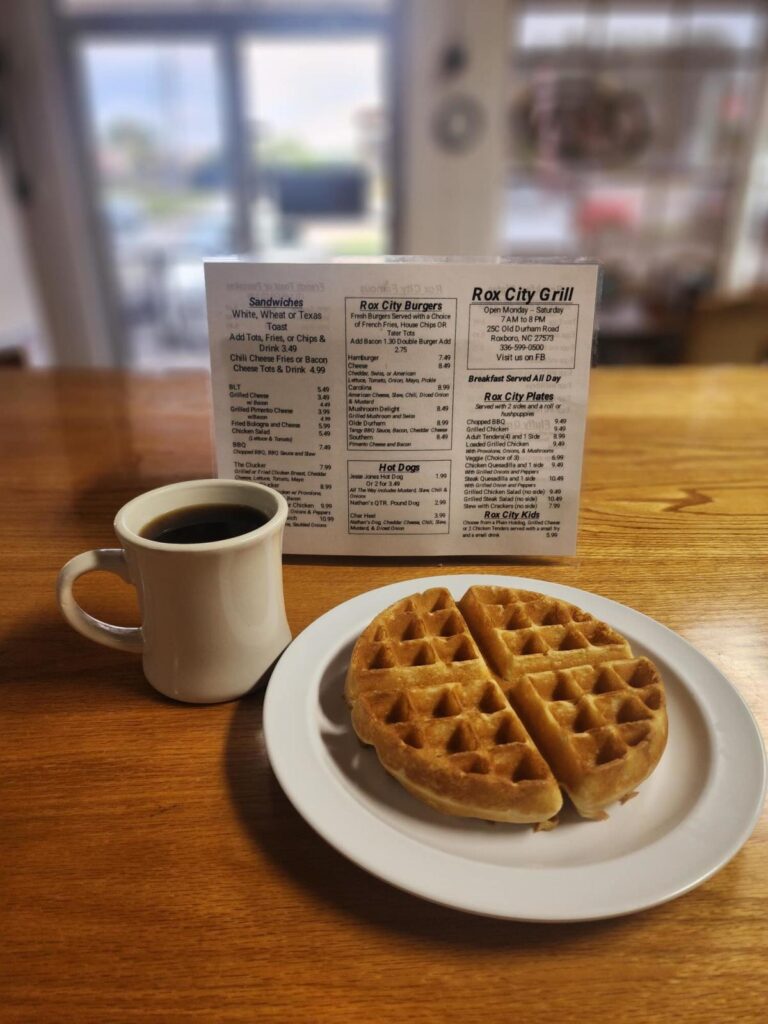 A white mug of coffee and a waffle on a white plate sit on a table in front of a diner menu