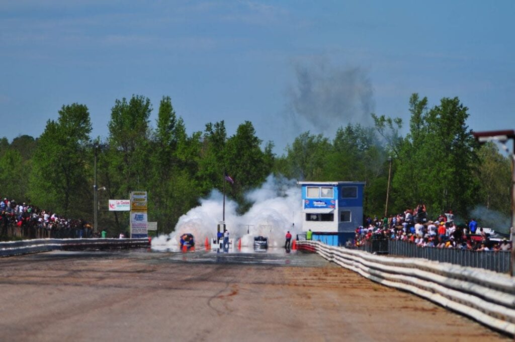 Two motor cars emitting plumes of white smoke at the start of a race