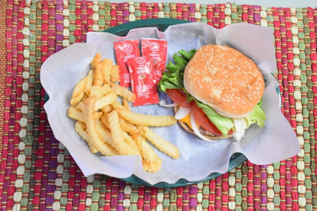 A cheeseburger and French fires with three packets of ketchup served in a green plastic basket on top of a colorful placemat