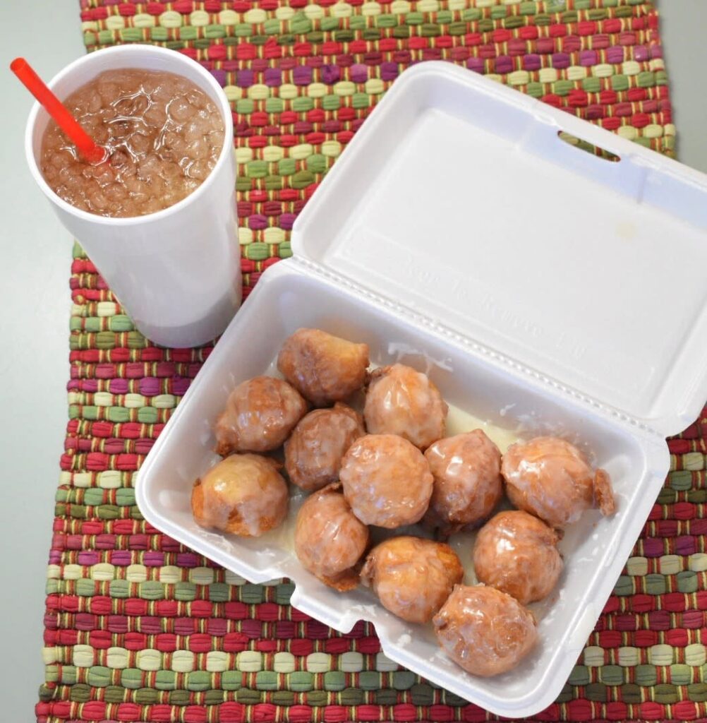 A white Styrofoam container with a dozen glazed donut holes and a Styrofoam beverage site on a colorful placemat