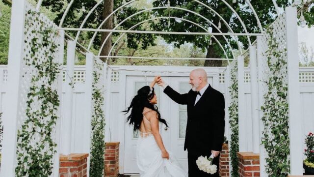 A groom wearing a black tuxedo and holding a bouquet of white flowers twirls a bride wearing a long white dress