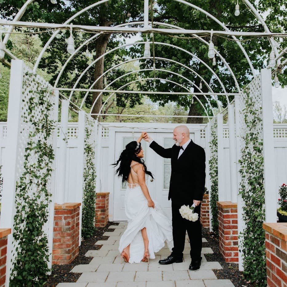 A groom wearing a black tuxedo and holding a bouquet of white flowers twirls a bride wearing a long white dress