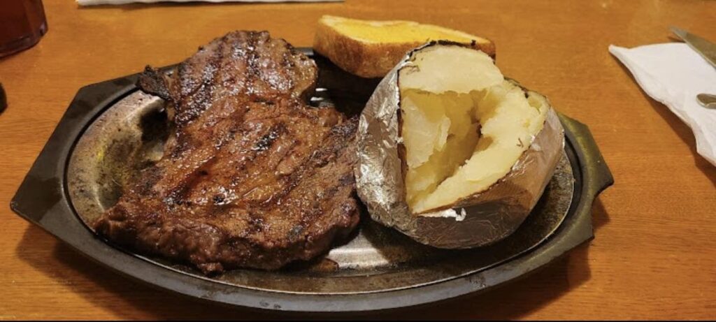 An oval-shaped metal plate with steak, a baked potato and slice of toast