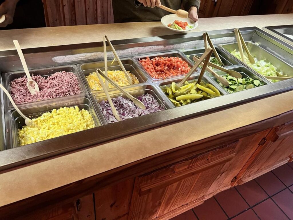 A buffet of salad toppings with serving tongs in each container