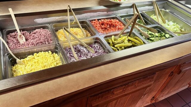 A buffet of salad toppings with serving tongs in each container