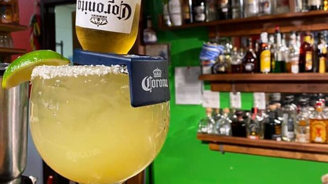 A margarita in a salt-rimmed glass sits on top of a wooden bar with a bottle of beer tipped into it