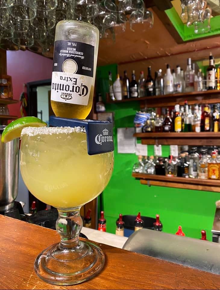 A margarita in a salt-rimmed glass sits on top of a wooden bar with a bottle of beer tipped into it