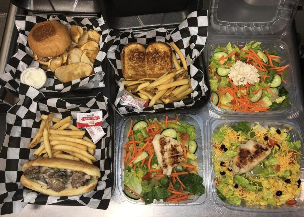 Plastic to-go containers with burgers, grilled cheese sandwiches, and salads