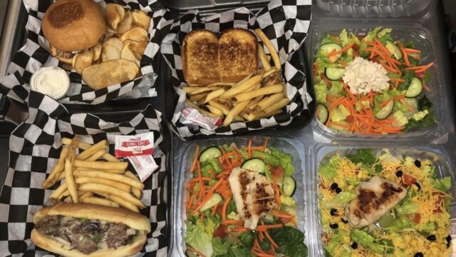 Plastic to-go containers with burgers, grilled cheese sandwiches, and salads